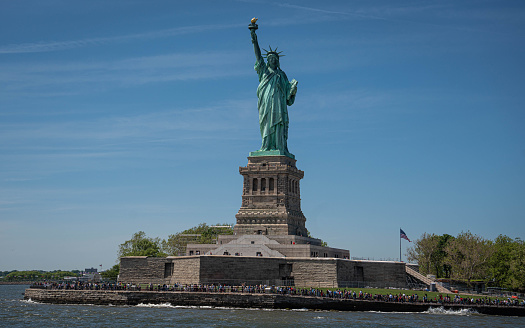 Statue of Liberty stands watch in New York Harbor on its stone pedestal against a blue sky.