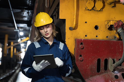 Female engineer using digital tablet for maintenance locomotive engine, wearing safety uniform, helmet and gloves in locomotive repair garage