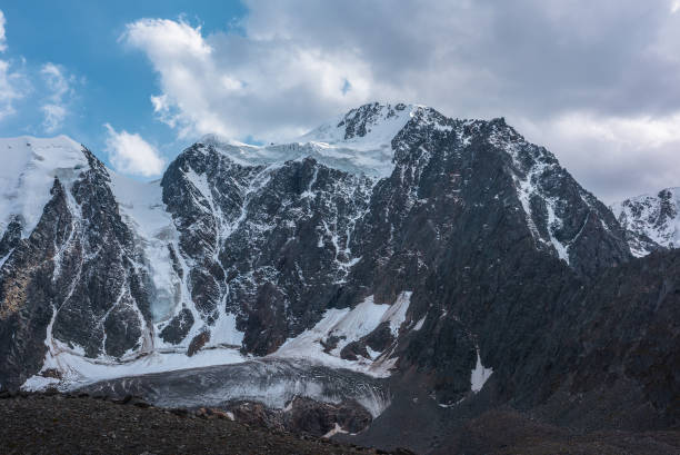 istock Atmospheric landscape with large snow mountain range under clouds in blue sky. Awesome high snowy mountains in changeable weather. Dramatic scenery with snow-covered mountain wall under cloudy sky. 1691408103
