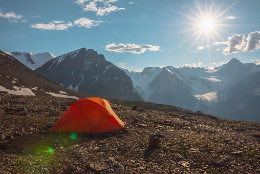 Awesome alpine landscape with orange tent with view to high snow mountains and large glacier. Bright sun in cloudy sky above snowy mountain range. Tent with top view to glacier tongue shining in sun.
