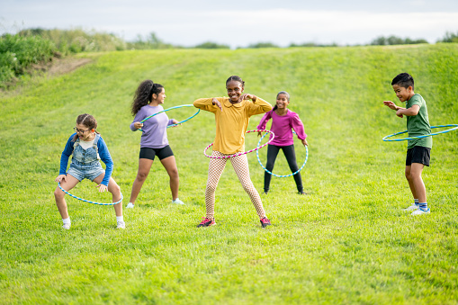 A small group of multi-ethnic school aged children are seen playing with Hula-Hoops during a recess break.  They are each dressed casually and are smiling as they focus on seeing who can spin their hoop the longest.