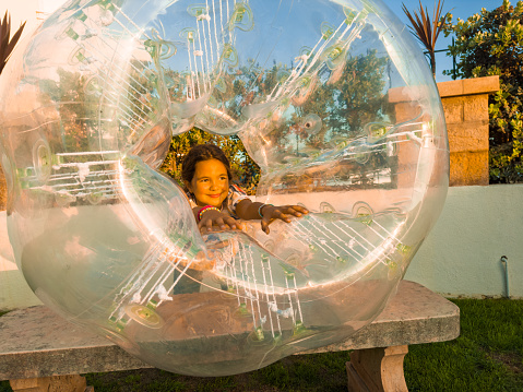 A horizontal photo of a girl and a boy standing outdoors playing with big bubbles.