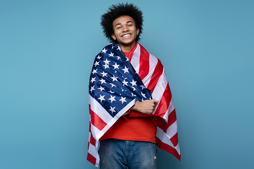 Confident smiling African American man covering with American flag isolated on blue background. Young emotional patriot celebration Independence day looking at camera. July 4th