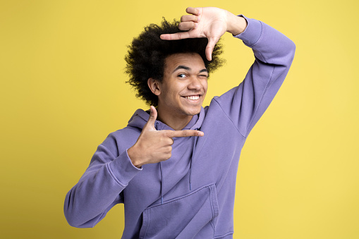 Young smiling African American photographer making frame with hands looking at camera isolated on yellow background