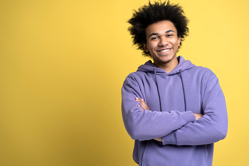 Handsome smiling African American man with arms crossed isolated on yellow background, copy space. Happy smart student wearing stylish purple hoodie looking at camera, studio shot. Education concept