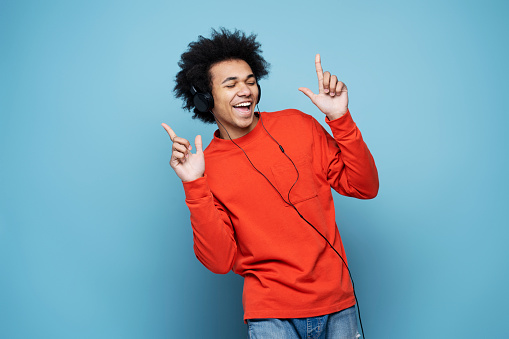 Happy emotional African American man wearing headphones listening music, singing song and dancing isolated on blue background. Technology, positive emotions concept