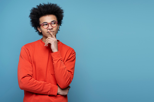 Young pensive African American student wearing eyeglasses holding finger near face, looking for creative solution isolated on blue background, copy space