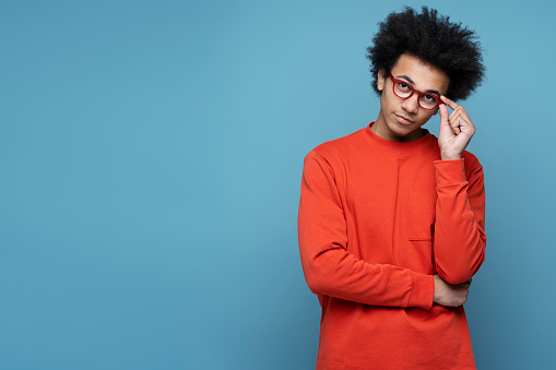 Portrait of smart pensive university student wearing stylish eyeglasses looking at camera isolated on blue background, copy space. Education concept
