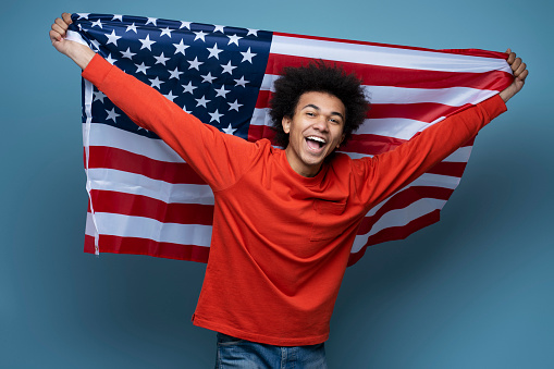 Young happy African American man holding American flag isolated on blue background. Emotional patriot celebration Independence day looking at camera