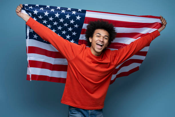 African American man holding American flag. Emotional patriot celebration Independence day Young happy African American man holding American flag isolated on blue background. Emotional patriot celebration Independence day looking at camera patriotic man stock pictures, royalty-free photos & images