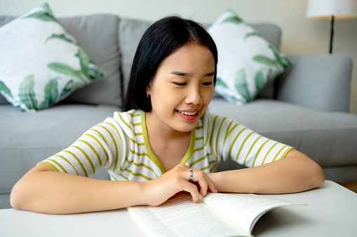 Portrait of Happy beautiful young Asian woman reading a book while relaxing and sitting on the floor in the living room at home. Leisure, hobby, lifestyle and people concepts.