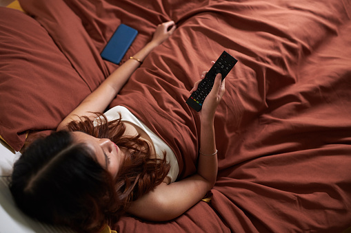 Young woman relaxing in bed in the evening and watching movie, view from above