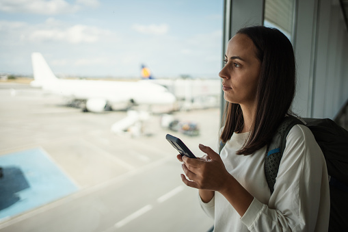 A woman stands by the window at the airport, using a phone while waiting for her departure.