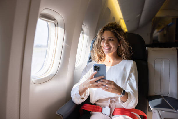 Female traveler using her cell phone in an airplane Latin American female traveler using her cell phone in an airplane and looking through the window first class stock pictures, royalty-free photos & images