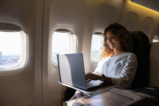 Happy Latin American female airplane passenger working on her laptop while traveling - travel concepts