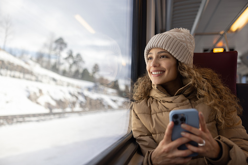 Portrait of a happy woman riding the train in the winter while using her phone and looking through the window