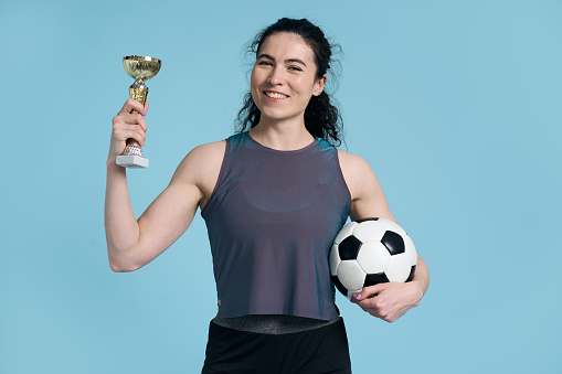 Studio portrait of skilled player soccer woman holding football ball and golden cup, celebrating winning the championship of her female team, smiling looking at camera, isolated on blue background