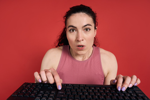 Close-up studio portrait of a Caucasian sportswoman typing text on computer PC keyboard, looking surprised, posing with her mouth open, on isolated red background. People and Technology. Laptop POV