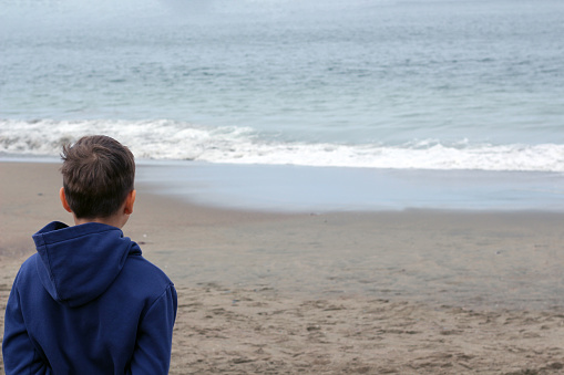 Portrait of a 10-year-old blond boy on the sandy beach on a cloudy day. The boy is dressed in a warm sweatshirt. He sits with his back to us.