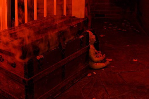 A zombie horror baby doll character holding a bloody brain in its lap is hiding behind an ancient, spooky Halloween night crypt-like steamer trunk coffin which is cracked open - allowing clouds of ominous, hazy mist and eerie apparitions to seep out - gleeful smoky ghosts finally escaping centuries trapped in suffocating captivity!