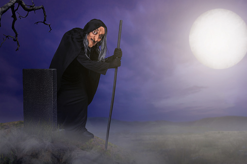 Old witch in a cloak with a stick standing in the graveyard with a night scene background. Scary witch Halloween concept