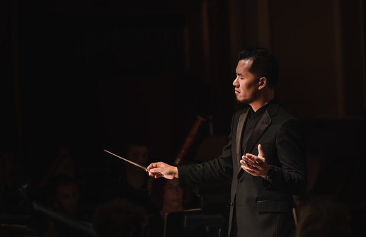 Middle aged Asian male conductor conducting orchestra in dark hall