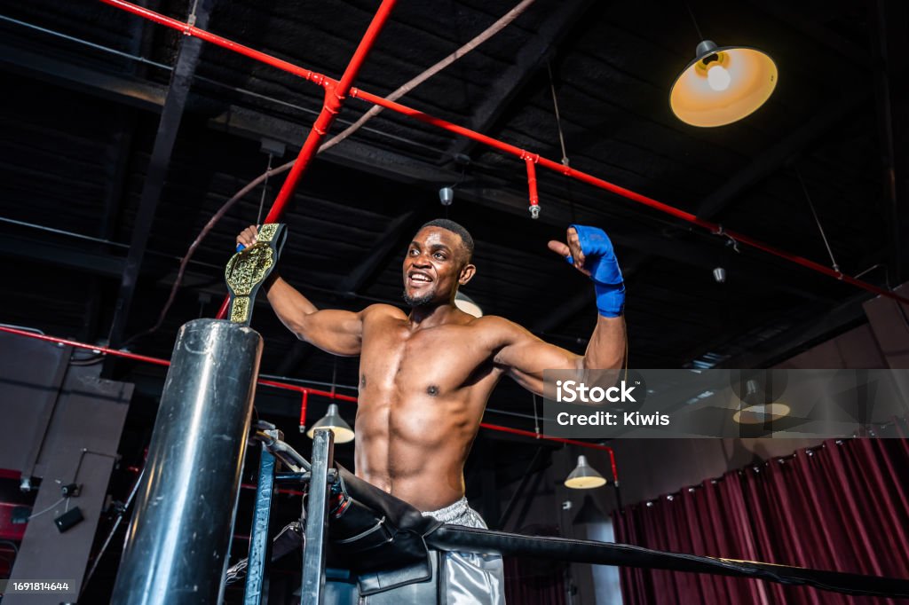 African muscular boxer raises his arms after win tournament on stage. Attractive male athlete fighters shirtless feel happy and excited, enjoy boxing exercise in the ring at stadium on competition. Wrestling Stock Photo
