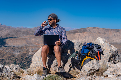 30-year-old model with a backpack. Wearing a hat and sunglasses. In a steep, mountainous area at high altitude. Open air. Mountain, nature view. Male model using a laptop. Getting his work done. Freelancer. Video calling is a multi-purpose topic.He has a hot water thermos and a backpack with him. Trekking at high altitudes. Exploration and adventure.He drinks something from the thermos next to him.
