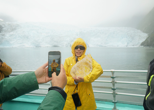 Tourist taking smartphone photos of a woman holding ice from an iceberg. Holgate Glacier in the rain. Kenai Fjords National Park. Seward. Alaska.