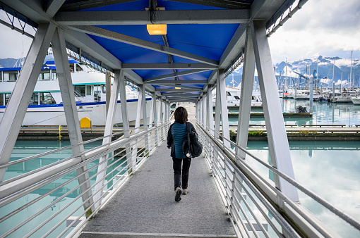 Woman with a backpack walking to the cruise boat. Sailboats and mountains in the distance. Seward. Alaska.