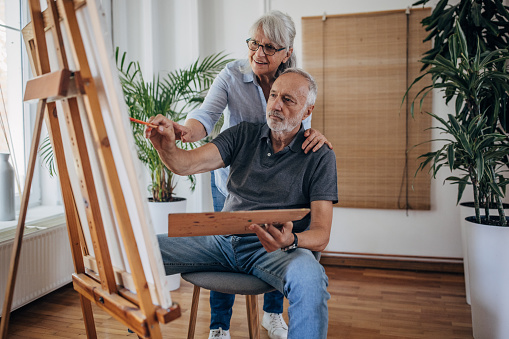 Senior couple at home, man is painting while his wife is assisting him in his work.