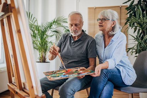 Senior couple at home, man is painting while his wife is next to him looking at his work.
