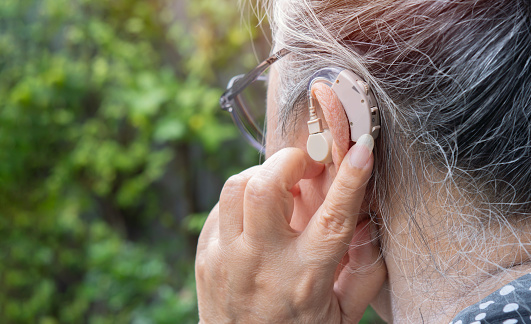 Closeup of a senior woman inserting and adjusting a hearing aid in her ear.  Selective focus on the hearing aid.