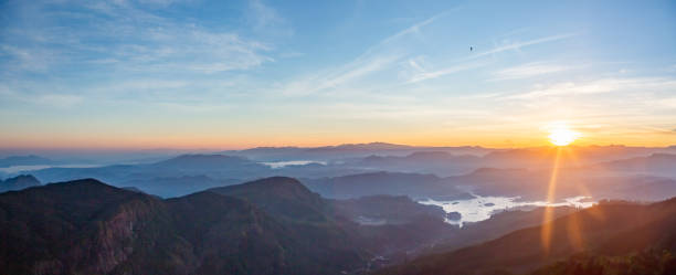 istock Panoramic View of a Sunrise/Sunset over a Mountain Range with Orange and Blue Sky, Green Mountains, and Clouds. 1691970795