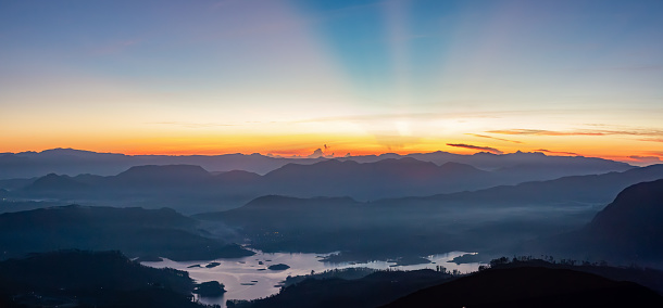 A panoramic view of a sunrise over a mountainous landscape. The sky is a gradient of orange and blue, with rays of sunlight shining through the clouds. In the foreground, there is a serene lake reflecting the vibrant colors of the sky. The landscape consists of multiple layers of mountains, creating a sense of depth and grandeur. The photo is taken from a high vantage point- Adam's peak in Sri Lanka, providing a sweeping view of the entire scene. The overall mood of the image is peaceful and tranquil.