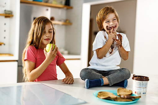 Small girl enjoying while eating an apple in the kitchen while her little brother is eating chocolate spread.