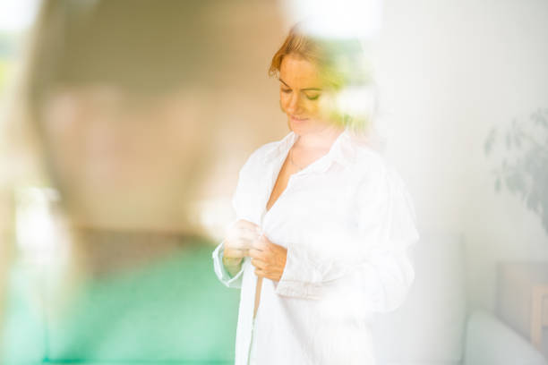 View through window of beautiful woman buttoning her white shirt at home