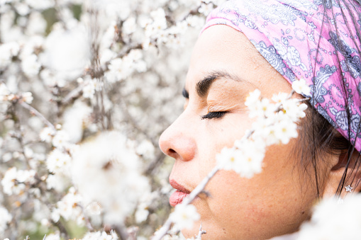 Close up profile shot of latin woman with closed eyes and headscarf smelling and enjoying the immensity of the almond blossom on a beautiful spring day