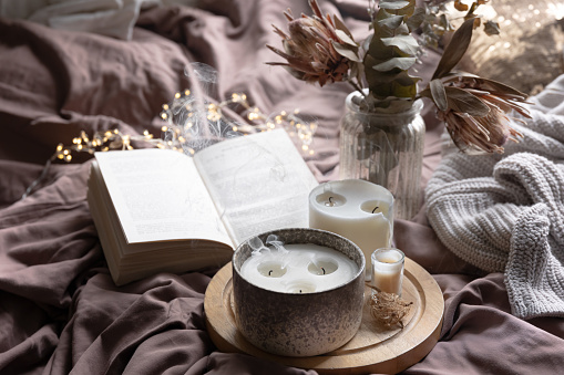 Cozy home composition with extinguished candles, book and knitted item in the bed, cozy home concept.