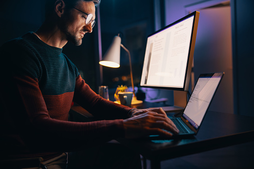 Caucasian man working late at night in his home office, engaged in a project using a laptop. Business man using technology to achieve after-hours productivity in remote work.