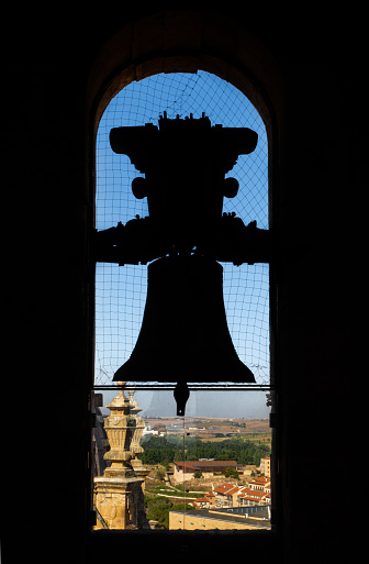 Backlit silhouette of the bronze bell of the old cathedral with the view of the city and mountains of Salamanca from the church tower. Protected with an anti-pigeon mesh.