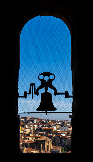 Backlit silhouette of bronze bell of the old cathedral with the view of the city of Salamanca from the church tower. Protected with an anti-pigeon mesh.