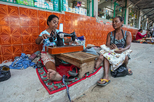 Toliara, Madagascar - May 01, 2019: Unknown older Malagasy woman sitting on ground at street next to market, mending clothes with hand crank sewing machines