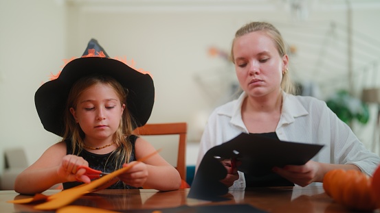 A small girl is wearing witch costume and making Halloween decorations with her mother in the living room at home.