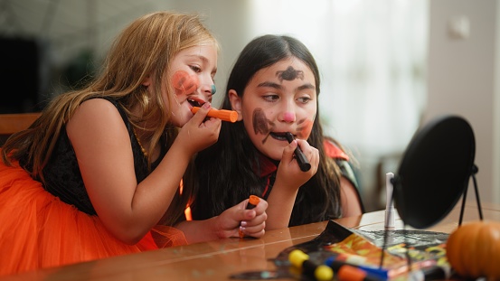 Two small female children friends are painting their faces for Halloween at home.