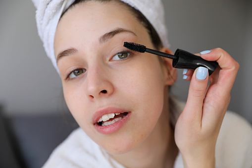 The girl paints her eyelashes. A young attractive woman in a white towel and bathrobe looks in the mirror in the bathroom and paints her eyelashes with mascara. Beautician, make-up concept at home. Close up view