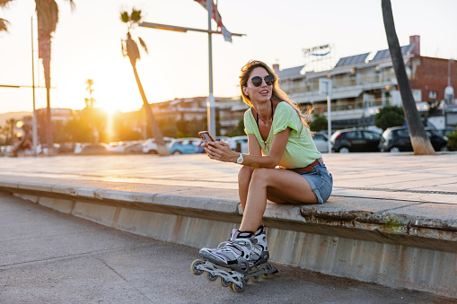 Cool young caucasian woman sitting on a concrete bench on the beach. She is holding a smartphone in her hands and has rollerblades on her feet. The sun is setting. The woman is wearing vibrant colourful summer clothes and sunglasses.