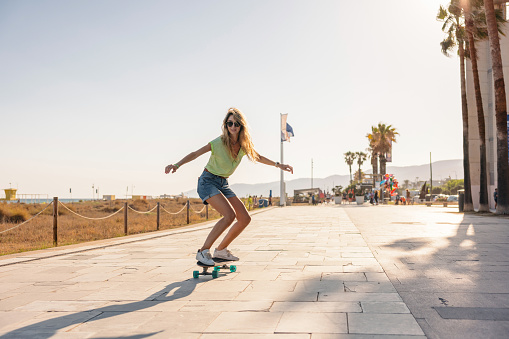 Cool sporty Caucasian woman enjoying her evening by riding a penny board on the boardwalk by the Barceloneta beach. She is having fun. The woman is attractive and is waring vibrant summer clothes and some sunglasses.