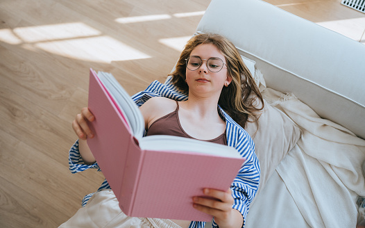 Teen girl laying on couch reading book at home. Focused schoolgirl In glasses holds book, remote learning. Distant education. Caucasian girl bored spending time with literature, wants walk outside.