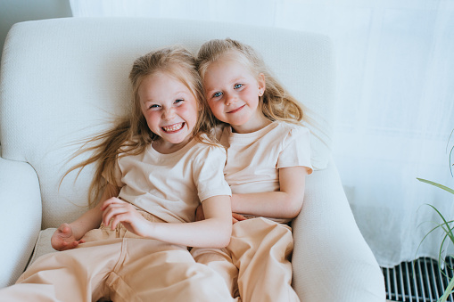 Two blonde Swedish little sisters sitting in chair looking at camera happily. Preschooler girls spending time home together. Childhood, domestic leisure. Sisterhood.